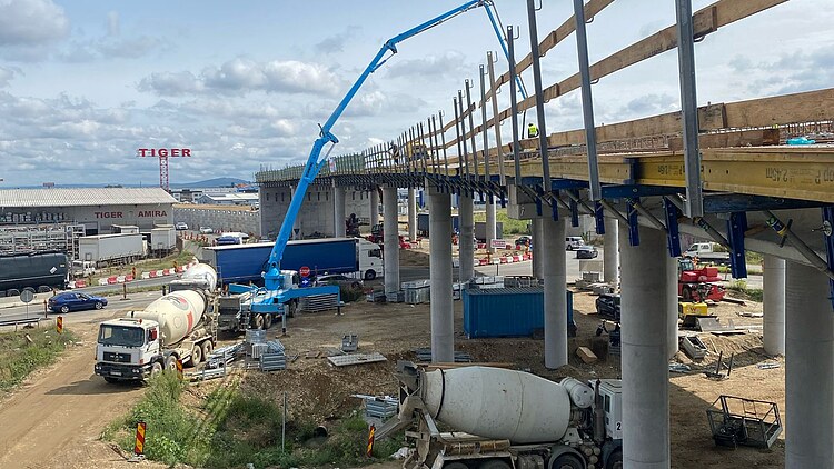 A concrete pump is delivering fresh concrete to a bridge under construction, complete with piers and formwork, whilst concrete mixers are delivering concrete to a building site near a road; buildings and traffic can be seen in the background.