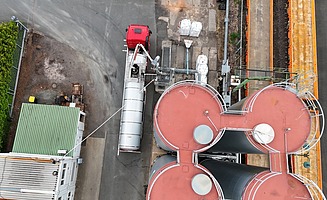 Aerial view of an industrial bitumen plant with several storage tanks, pipelines and a loading station where a tanker lorry is being filled.