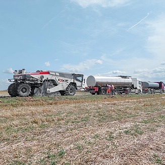 Spezialmaschine für Kaltrecycling in situ verarbeitet vorhandenes Straßenmaterial direkt vor Ort, während Tankfahrzeuge Bindemittel zuführen und Personen die Arbeiten begleiten