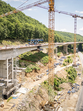 Brückenbaustelle mit hohen Betonpfeilern, im Bau befindlichem Überbau und mehreren Turmdrehkränen, umgeben von Wald und Geländeeinschnitten.