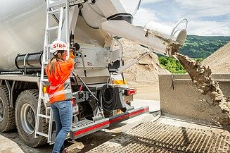 A person wearing protective clothing operates a truck mixer whilst fresh concrete is being discharged via a chute into a collection system on a building site.