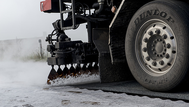 Spray bar of a road construction vehicle evenly distributes bitumen emulsion on an asphalt surface to create bonding bridges and layer bonding; visible are nozzles that apply the emulsion and a vehicle wheel in the foreground.