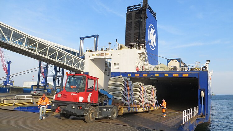 Loading of prefabricated concrete segments onto a RoRo ship in the harbour via a ramp; a heavy goods vehicle transports the elements, accompanied by people in protective clothing
