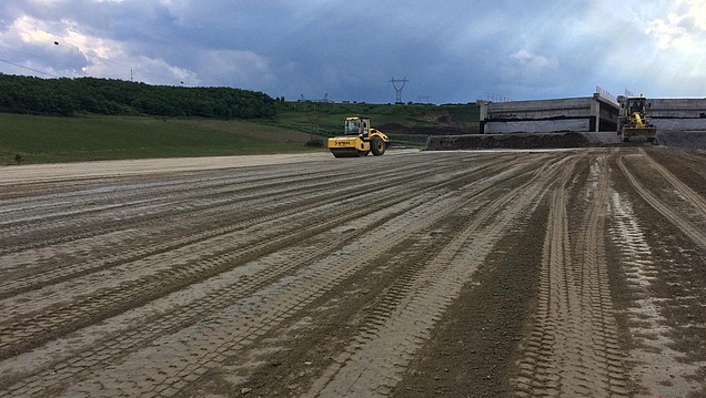 Walzenzug verdichtet eine großflächige Erd- oder Baustoffschicht auf einer offenen Baustelle mit Baumaschinen und einer Betonbaukonstruktion in ländlicher Umgebung.