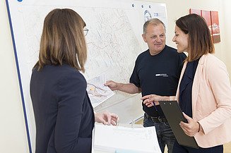 Drei Personen stehen vor einer großformatigen technischen Karte an einer Wandtafel; eine Person hält einen ausgedruckten Plan, eine weitere hält ein Klemmbrett, während gemeinsam auf den Plan verwiesen wird.