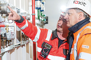 Two people in protective clothing and helmets check measuring displays and fittings on a technical system; one person points to an analogue measuring device, another holds a digital measuring instrument in their hand - the operation and quality assurance of a mobile concrete mixing plant is shown.