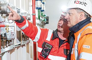 Two people in protective clothing and helmets check measuring displays and fittings on a technical system; one person points to an analogue measuring device, another holds a digital measuring instrument in their hand - the operation and quality assurance of a mobile concrete mixing plant is shown.
