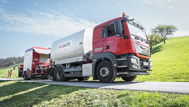 A tanker with a spraying system for bitumen emulsions is carrying out a surface treatment on an asphalt road; a downstream spreader and a rural setting with meadows and trees can be seen in the background.