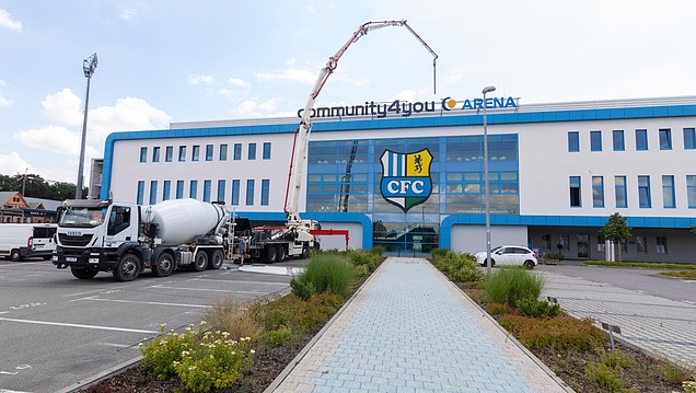 A concrete mixer and concrete pump in action outside a public building bearing the inscription ‘Community4you Arena’; concrete is being laid on a building site