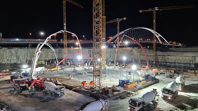 A large industrial and commercial construction site at night, with several concrete pumps, truck mixers and cranes pouring concrete into a reinforced concrete structure