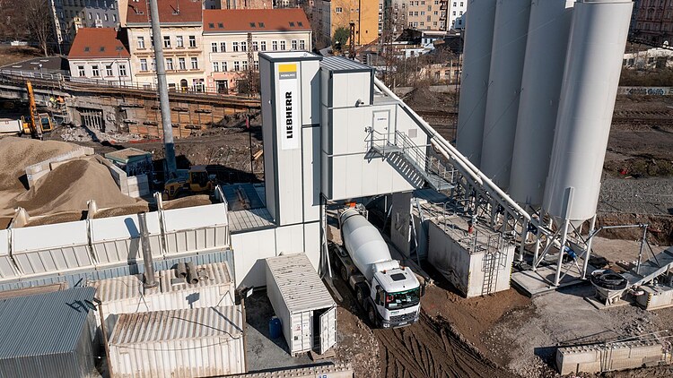A mobile concrete batching plant with silos and conveyor systems on a building site situated between residential buildings; a concrete mixer truck is waiting to be loaded at the plant module.