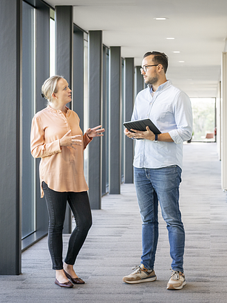 Two people in conversation in a modern office corridor with large windows; one person holds a tablet while discussing content together - collaboration in everyday working life.