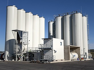 Industrial bitumen plant with several high, cylindrical metal storage tanks, connected by pipelines and supplemented by technical ancillary buildings on a paved site.