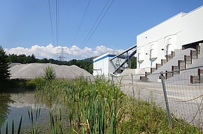 Exterior view of a former concrete and building materials plant with conveyor technology, storage areas and bulk material next to a fenced-in area and a small body of water; the picture documents the condition of the plant as part of the dismantling and documentation of a mobile concrete mixing plant.