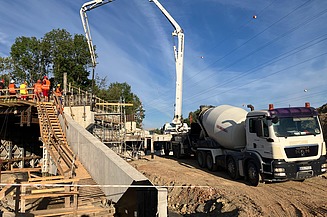 A concrete mixer truck and a concrete pump are delivering and pumping fresh concrete onto a construction site, whilst several people on a platform are preparing for the pour; trees and overhead power lines can be seen in the background.