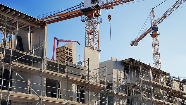 Mehrgeschossiger Rohbau mit Gerüsten und Mauerelementen, mehrere Turmdrehkräne heben Baumaterial auf einer Hochbaustelle.