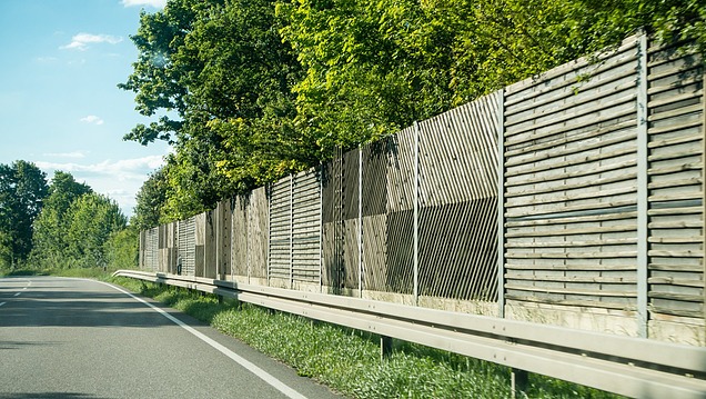 Road with lateral noise barrier made of wooden and structural elements along the edge of the carriageway, surrounded by trees and green spaces