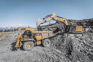 A hydraulic excavator is loading rock material into a tipper lorry on a spoil heap containing loose bulk material in a mining or processing area.
