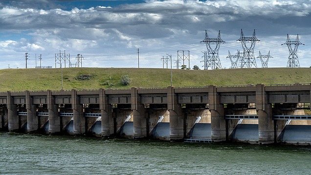 A weir with several concrete piers and open weir gates on a river; electricity pylons and a raised embankment are visible in the background.