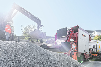 Excavator loads aggregate into a road construction machine while people in protective clothing operate the process