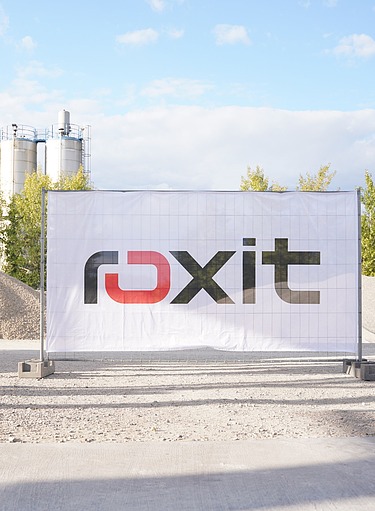 Construction fence with Roxit logo in front of gravel heaps and a concrete mixing plant with silos, surrounded by trees under a blue sky.