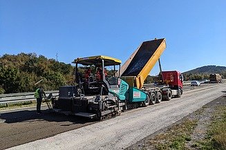 Road construction machine applies material to a carriageway while a lorry tips the material and personnel accompany the work on a motorway construction site.