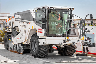 Wirtgen road milling machine with large wheels and milling unit on a company site; the driver's cab, hydraulic components and attachments are visible - part of the company's own machine fleet for road maintenance.