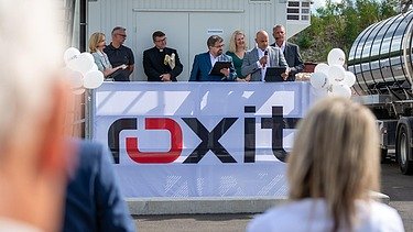 Festveranstaltung vor einer roxit-Anlage am Standort Planá: Mehrere Personen stehen auf einer Bühne und sprechen zu den Gästen. Im Vordergrund ist ein großes roxit-Banner zu sehen.