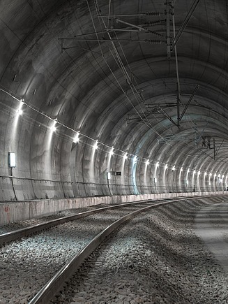Eisenbahntunnel mit verlegten Gleisen auf Schotterbett, Betonwänden, Beleuchtung und installierter Oberleitung entlang der gesamten Tunnelröhre.