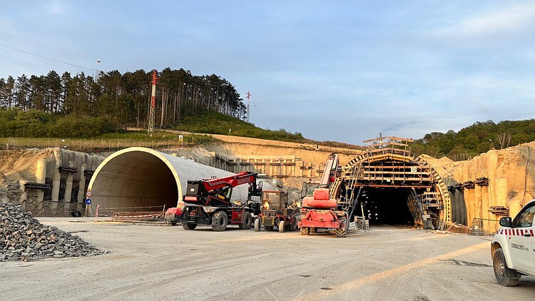 A tunnel construction site with two tunnel portals, one lined with precast concrete elements and the other under construction using timber formwork; in front of them, construction machinery and vehicles are parked on an open construction site.