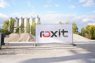 Exterior view of an industrial site with storage tanks, stockpiles of bulk materials and a banner with the company logo, surrounded by trees on a paved factory site.