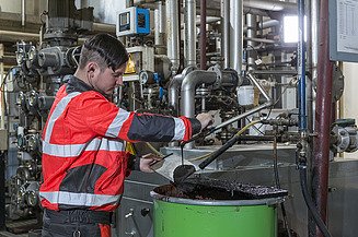 Person takes a sample of bitumen emulsion from an industrial production plant with pipework and measuring equipment