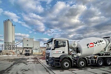 Truck mixer on the site of a concrete mixing plant with silos and production buildings under a cloudy sky