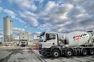 A concrete mixer on the site of a concrete batching plant, with silos and production buildings, under a cloudy sky