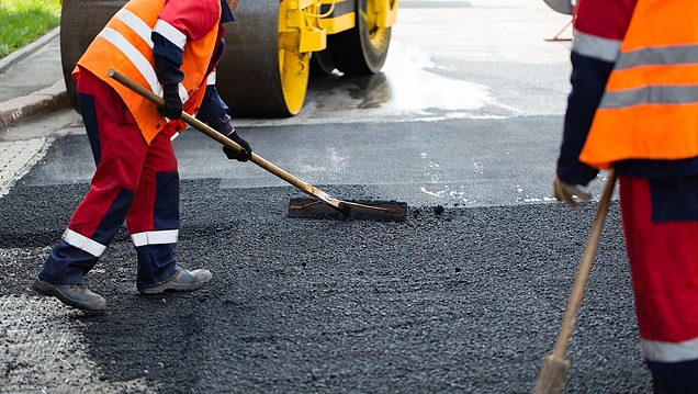 Straßenbauarbeiten mit frischem Asphalt, der von einer Person mit einem Handwerkzeug verteilt wird, während im Hintergrund eine Walze die Oberfläche verdichtet