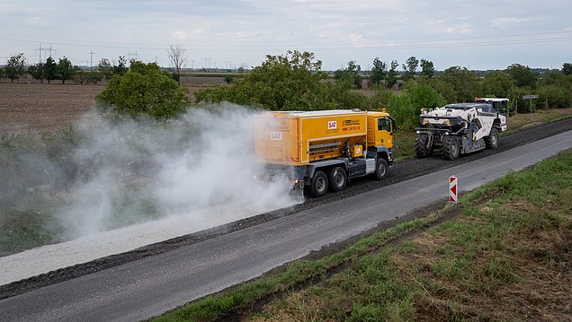 Spezialfahrzeug sprüht Material auf eine Fahrbahn, gefolgt von einer Straßenbaumaschine auf einer ländlichen Straße; sichtbar ist eine Staub- oder Nebelentwicklung während der Bearbeitung
