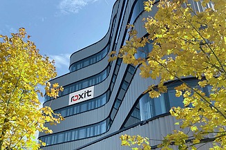 A modern office façade with curved lines and the roxit logo, surrounded by yellow-coloured trees against a blue sky.