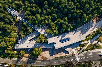 Aerial view of a modern building complex with green roof surfaces, embedded in a dense forest landscape next to a road.