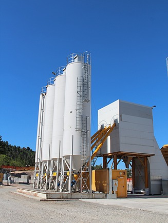 Several high concrete silos with ladders and railings next to a mobile concrete mixing plant with yellow conveyor pipes on an open construction site area under a blue sky.