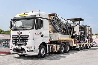 Schwertransport mit Lkw und Tieflader transportiert eine Straßenbaumaschine, darunter Fräse oder Spezialgerät, im Rahmen des Maschinenparks für die Straßeninstandhaltung; deutlich sichtbar sind Ladefläche, Hydraulikarm und technische Komponenten.
