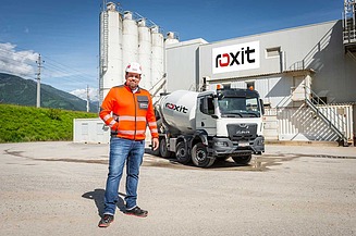 Mitarbeiter in Warnschutzkleidung steht vor einem Betonwerk mit roxit-Logo, daneben ein Fahrmischer auf dem Werksgelände.