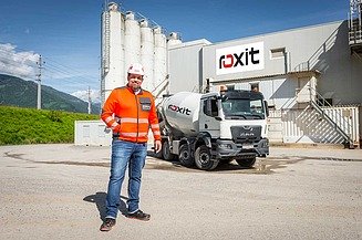 Employees in high-visibility clothing stand in front of a concrete plant with the roxit logo, next to a truck mixer on the factory premises.