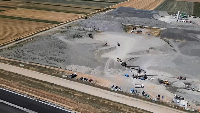 Luftaufnahme einer großflächigen Baustoffanlage mit Materialhalden, Förderanlagen und Baumaschinen in einer offenen Landschaft