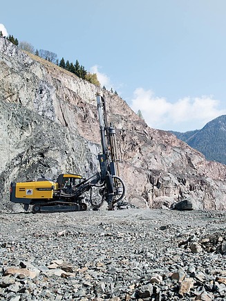 A drilling rig is operating on a rock face in a quarry, surrounded by rubble and mountainous countryside.