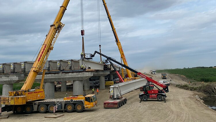 Two large mobile cranes are lifting a precast concrete element onto a bridge structure, whilst several people are supervising the positioning of the element on a motorway construction site; vehicles and unpaved tracks are visible in the background.