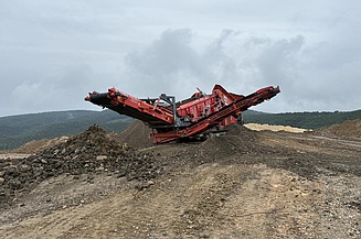 Mobile Brech- und Siebanlage verarbeitet Gesteins- und Recyclingmaterial auf einer offenen Fläche, umgeben von Halden und hügeliger Landschaft.