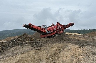 Mobile Brech- und Siebanlage verarbeitet Gesteins- und Recyclingmaterial auf einer offenen Fläche, umgeben von Halden und hügeliger Landschaft.
