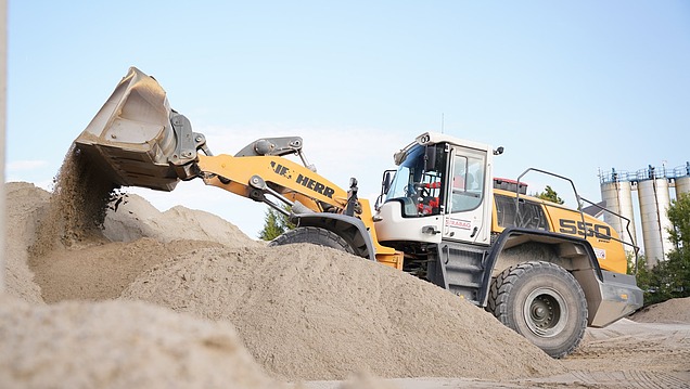 Radlader schüttet Sandmaterial auf einer Baustoffanlage um, im Hintergrund sind Silos und technische Anlagen sichtbar.
