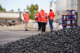 Kieshaufen im Vordergrund einer Baustelle, im Hintergrund stehen mehrere Personen in Warnkleidung neben einem Lkw und gelagerten Baustoffen.