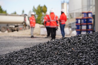 A pile of gravel in the foreground of a building site; in the background, several people in high-visibility clothing are standing next to a lorry and piles of building materials.